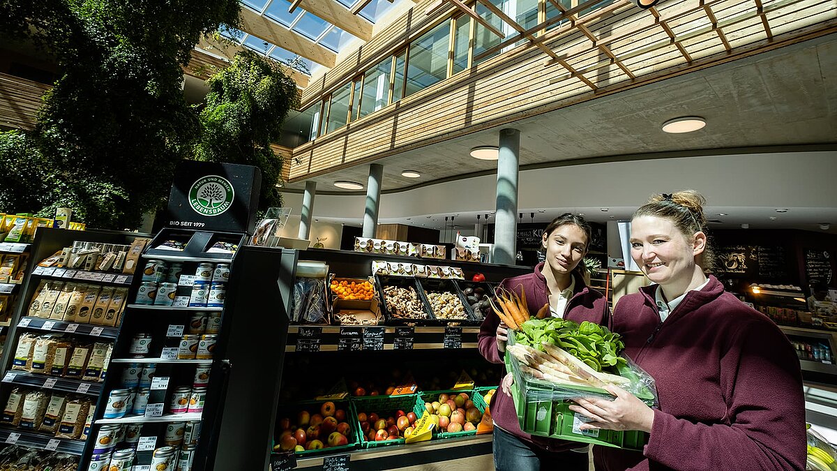 Zwei Mitarbeiterinnen im Naturkostladen Byodo in Mühldorf, präsentieren frisches Bio-Gemüse vor einem modernen, lichtdurchfluteten Verkaufsbereich mit Holz- und Glasakzenten.