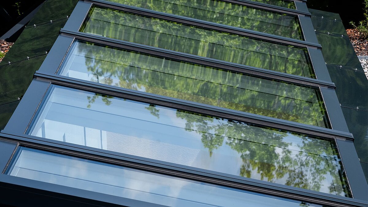 Nahaufnahme eines modularen Glasdachs von LAMILUX in einem Wohnhaus in Freiburg, mit Spiegelung von Bäumen in den Glasscheiben.